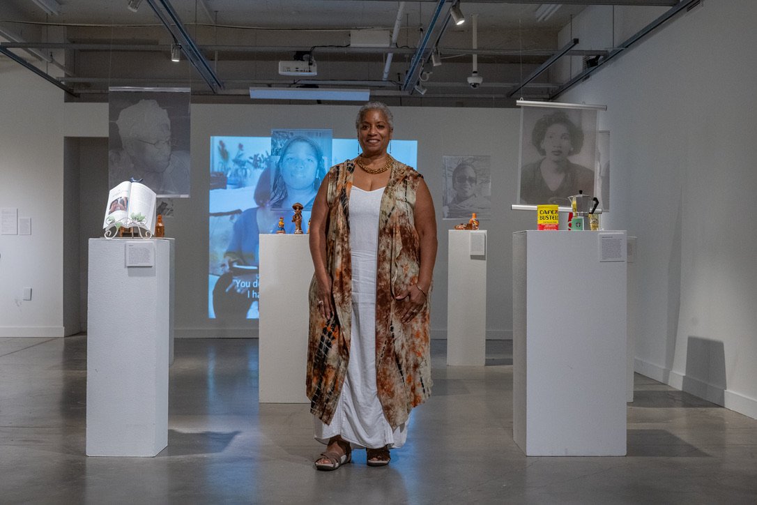 BasSheva Mañjon standing among artwork in a gallery, wearing a colorful sleeveless jacket over a white dress.