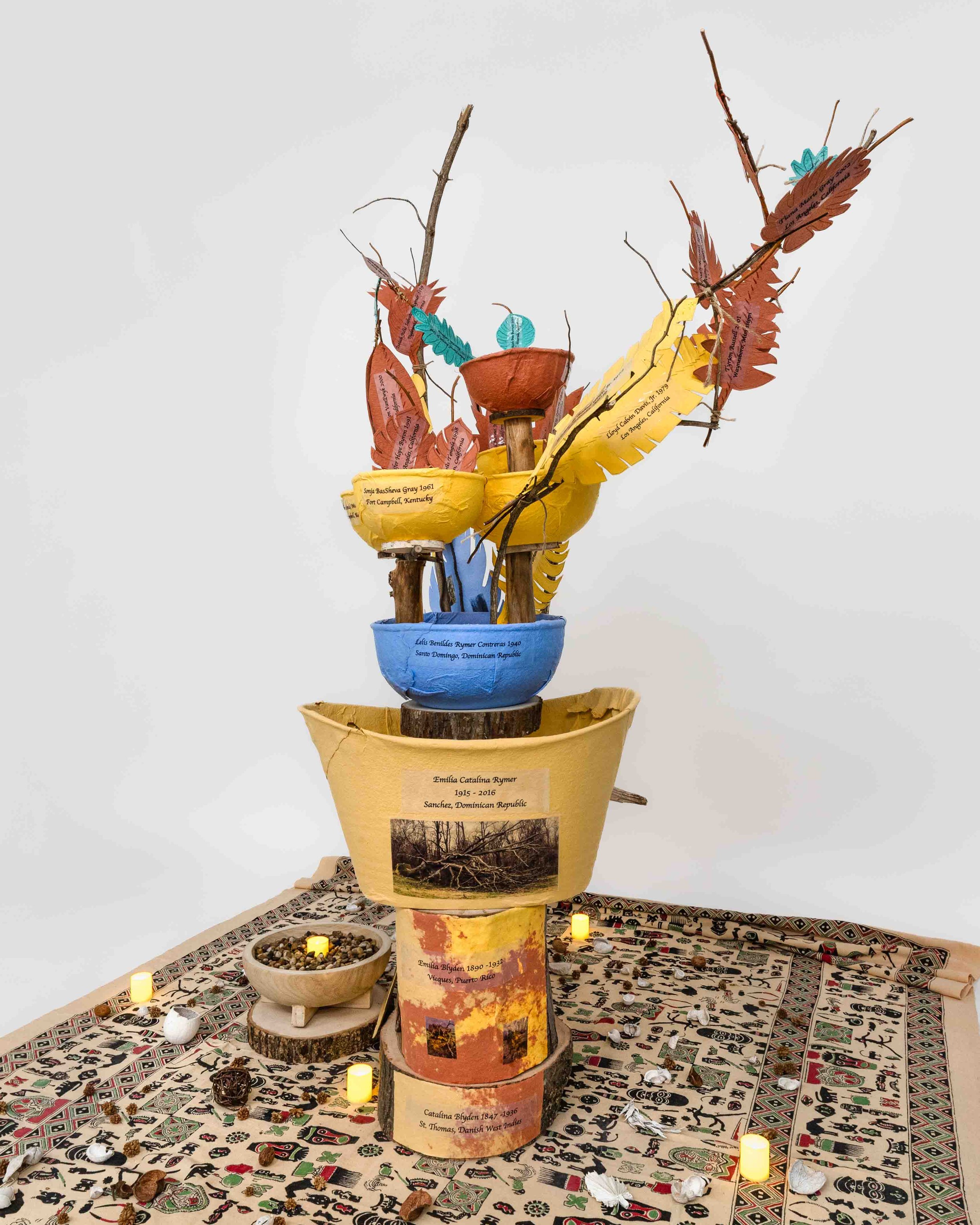 Installation of brightly colored handmade paper bowls, leaves, family photographs, and branches.